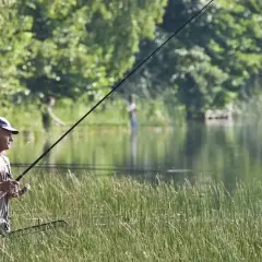 Mężczyzna z wędką w ręku, otoczony wysoką trawą, cieszy się spokojnymi godzinami brania ryb nad jeziorem.