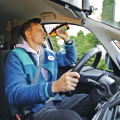 Mężczyzna za kierownicą pije piwo. Czy pasażer może pić alkohol? To niebezpieczne zachowanie.