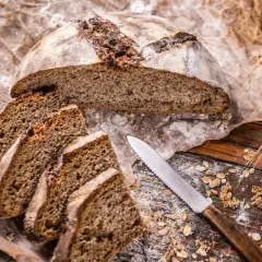 Chleb żytni, pokrojony w kromki, leży na papierze. Obok nóż i kłosy zbóż. Zastanawiasz się, jak długo piec chleb?
