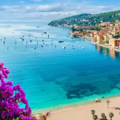 Blick auf einen malerischen Strand in Frankreich mit türkisfarbenem Wasser, Segelbooten und einer Küstenstadt, umrahmt von leuchtend lila Bougainvillea.