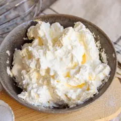 Schüssel mit luftiger, weißer italienischer Buttercreme, bereit für süße Kreationen. Daneben Zucker und ein Schneebesen.