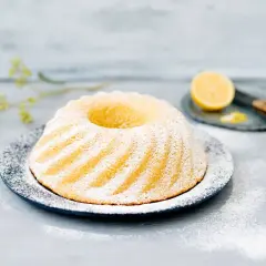 En vackert pudrad sockerkaka med citron, serverad på en mörk tallrik med en citronskiva och en kniv i bakgrunden.