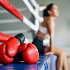 Red boxing gloves rest on the edge of a ring, hinting at a boxer puncher style. A woman sits in the background, her face turned away.