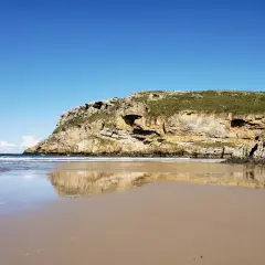 Reflejos dorados en la arena mojada de la playa de Fuentes, con acantilados cubiertos de verde bajo un cielo azul.