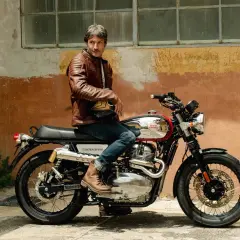 Uomo con giacca in pelle marrone seduto su una moto Royal Enfield Interceptor. I terminali moto cromati brillano sotto il sole.