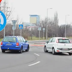 Niebieski Volkswagen i biały Fiat na rondzie. Kierowcy zastanawiają się, który zjazd z ronda na który pas wybrać, by bezpiecznie kontynuować jazdę.