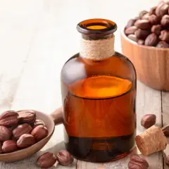 Bursztynowa buteleczka z płynnym złotem – czysty olej jojoba na włosy. Obok drewniana łyżka i miseczka pełna nasion jojoby.