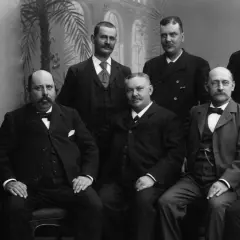 Gruppbild på män i 1900 talet kläder. De bär kostymer och flugor, redo för ett formellt porträtt.