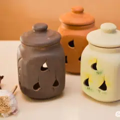 Drei Keramikbehälter in verschiedenen Farben mit Lüftungslöchern, daneben frischer Knoblauch und Zwiebeln. Perfekt für knoblauch haltbar machen.