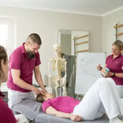 Ein Mann erklärt einer liegenden Frau eine physiotherapeutische Behandlung. Ein Skelett und eine Tafel mit anatomischen Zeichnungen verdeutlichen den Unterschied zwischen Ergo- und Physio-Therapie.