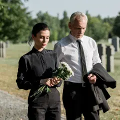 Smutna kobieta w czarnym stroju na pogrzeb trzyma kwiaty, obok mężczyzna w garniturze.