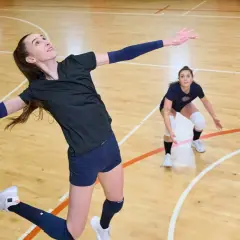 Kobieta w skoku do ataku w siatkówce, w tle dwie inne osoby.