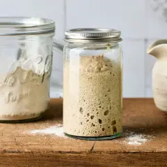 Ein Glas mit blubberndem Sauerteig, bereit zum Backen. Daneben ein Glas mit Mehl, um den Sauerteig anzusetzen.
