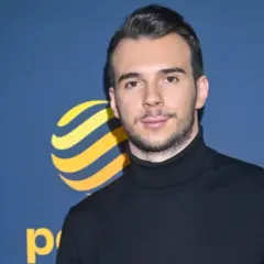 Młody mężczyzna, Albert Kosiński, w czarnym golfie na tle niebieskiej ścianki z logo.