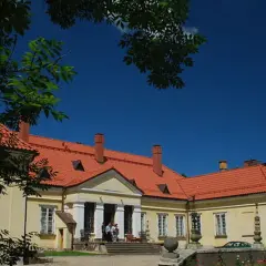 Pałac w Waplewie z czerwonym dachem i żółtymi ścianami, otoczony zielenią, pod błękitnym niebem.