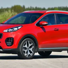 Czerwony Kia Sportage IV z kierowcą w okularach przeciwsłonecznych jedzie po drodze, otoczony zielenią.