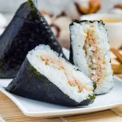 Three triangular onigiri triangle sushi with tuna filling, wrapped in nori seaweed, sit on a white plate with chopsticks and a cup of tea in the background.