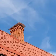 Ceglany komin na dachu pokrytym pomarańczową dachówką. Opierzenie komina wykonane z blachy trapezowej.