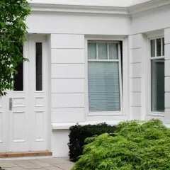 Białe drzwi i okna eleganckiego domu, otoczone bujną zielenią. Drzewo z liśćmi rzuca cień na fasadę, a krzewy dodają uroku.