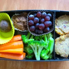 A bento box packed with bento nutrition: a date ball, muffin, grapes, carrots, broccoli, and two savory patties.