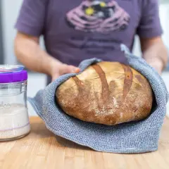 Un panadero muestra un pan artesanal recién horneado junto a un frasco de masa madre activa, listo para empezar a hacer masa madre.