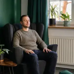 Mężczyzna w fotelu z zamkniętymi oczami, być może doświadcza objawów nerwicy. W tle zielona ściana, rośliny i okno.