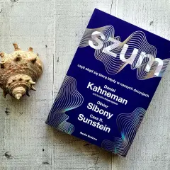 Książki Daniela Kahnemana, "Szum", obok muszli na drewnianym tle.
