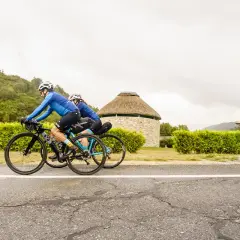 Ciclistas pedalean por la ruta EuroVelo 3, pasando junto a un edificio redondo con techo de paja y una estatua de un peregrino.