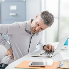 Mężczyzna z bólem pleców przy biurku, pracujący na laptopie. To może być objaw choroby zawodowej.