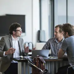 Three men discuss performance review tips for employees in a modern office setting.