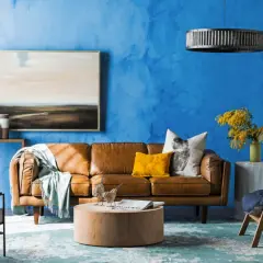 Ein Wohnzimmer mit leuchtend blauer Wand. Ein braunes Ledersofa, ein gelbes Kissen und ein Bild mit Landschaftsmotiv. Welche Farbe passt gut zu Blau? Hier: Gelb und Braun.
