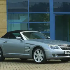 Srebrny Chrysler Crossfire auto z czarnym dachem stoi przed budynkiem z żółtej cegły i niebieskimi oknami.