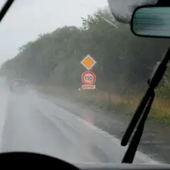 Conduire sous la pluie sur une route mouillée, avec des essuie-glaces en action et un panneau de rappel de vitesse 110.