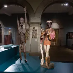 Two striking sculptures, one a wooden figure with raised arms, the other a painted figure holding a chalice, are displayed in a museum gallery, showcasing medieval African art.