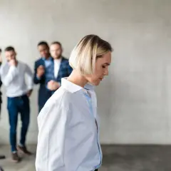 Smutna kobieta odwrócona tyłem, podczas gdy trzej mężczyźni za nią szepczą i wskazują na nią. To przykład mobbingu w pracy.