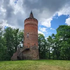 Stara, ceglana wieża zamkowa z kamiennym fundamentem, otoczona zielenią drzew, pod błękitnym niebem z chmurami.