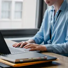 Osoba w niebieskiej koszuli pracuje przy laptopie, tworząc arkusz xlsx. Na biurku leżą notatniki i teczka.