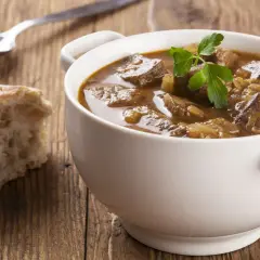 Aromaticzny gulasz wołowy w sosie własnym, podany w białej miseczce z gałązką natki pietruszki, obok kromki chleba i łyżki.