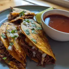 Tre saftiga birria tacos med strimlat kött, lök och koriander, serverade med en skål consommé och en limeklyfta.