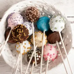 Kosz pełen kolorowych cake popsów: różowych, białych, czekoladowych i niebieskich, ozdobionych posypkami i kuleczkami.