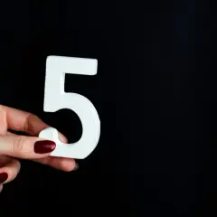 Dłoń z ciemnoczerwonym lakierem trzyma białą cyfrę 5, symbolizującą numerologię 5.