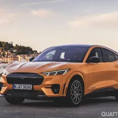 Arancione Ford Mustang Mach-E, una delle auto elettriche sportive più desiderate, parcheggiata con una pittoresca città costiera sullo sfondo.