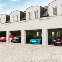 Piotr Śledź w czerwonej bluzie i białych spodniach stoi przy luksusowych samochodach: czerwonym Rolls-Royce, czarnym Rolls-Royce, niebieskim Lamborghini i czerwonym Bentleyu.