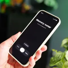 Dłoń trzyma smartfon z ekranem blokady, na którym widnieje "Nieznany numer" i numer telefonu. W tle zielone rośliny.