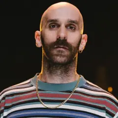 Łysy mężczyzna z zarostem, w kolorowej bluzie i złotym łańcuszku, wygląda jakby miał zaraz zacząć śpiewać "x ambassadors unsteady tekst".