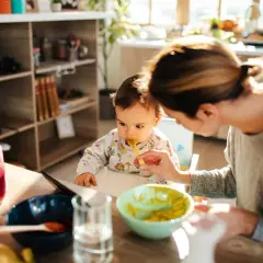 Mama karmi dziecko łyżeczką. Na stole są owoce i warzywa, idealne dla niemowlaka, który zaczyna poznawać, co może jeść 6 msc dziecko.
