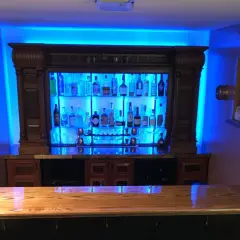 A stunning home bar setup, perfect for entertaining. This image shows how to build a bar with a wooden countertop, a backlit liquor cabinet filled with bottles, and vibrant blue ambient lighting.