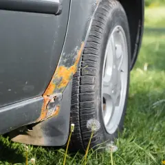 Widoczna rdza pod lakierem na progu samochodu, obok opony i zielonej trawy.