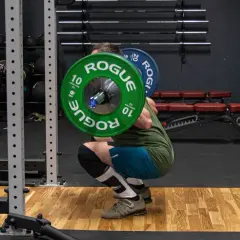Mężczyzna wykonuje przysiady ze sztangą na karku w siłowni. Zielone talerze "Rogue" ważą 10 kg każdy.