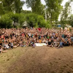 Große Gruppe feiert beim auerworld festival. Viele Menschen mit erhobenen Armen im Freien, umgeben von Bäumen und bunten Bannern.